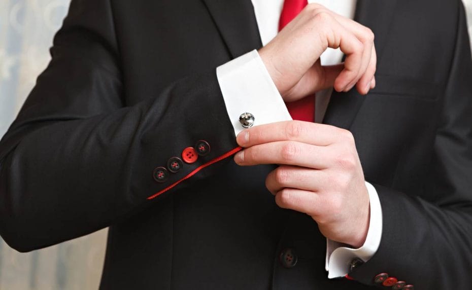 Man clasps cufflink on his shirt, wearing a black blazer and red tie.