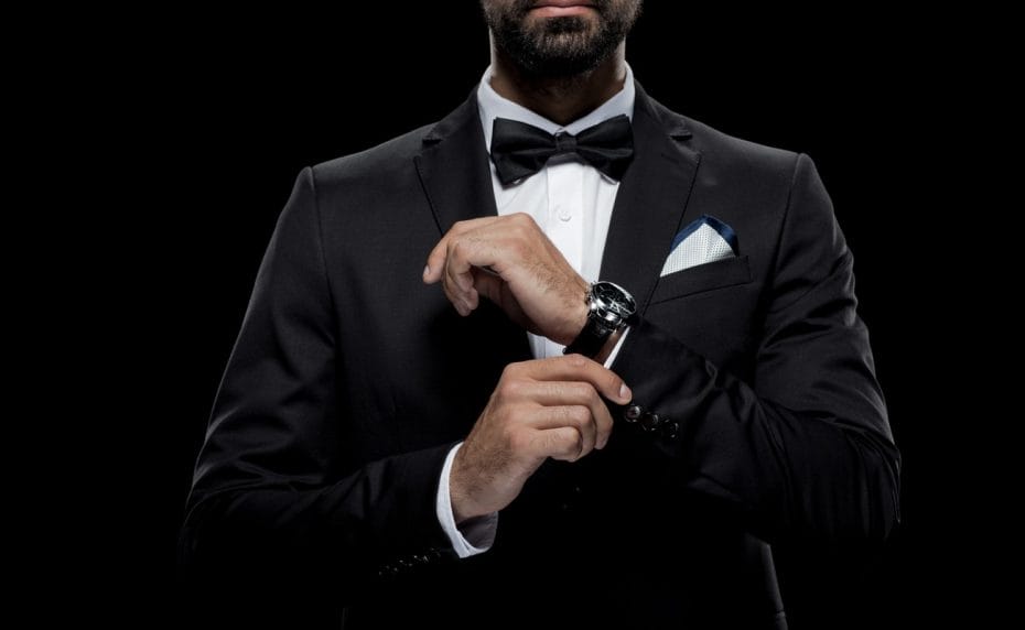 Man in a tuxedo with a bow tie and wristwatch, with a black background. 