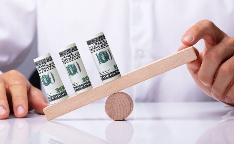 A person's hand balancing 3 rolled up dollar bills on a wooden seesaw.