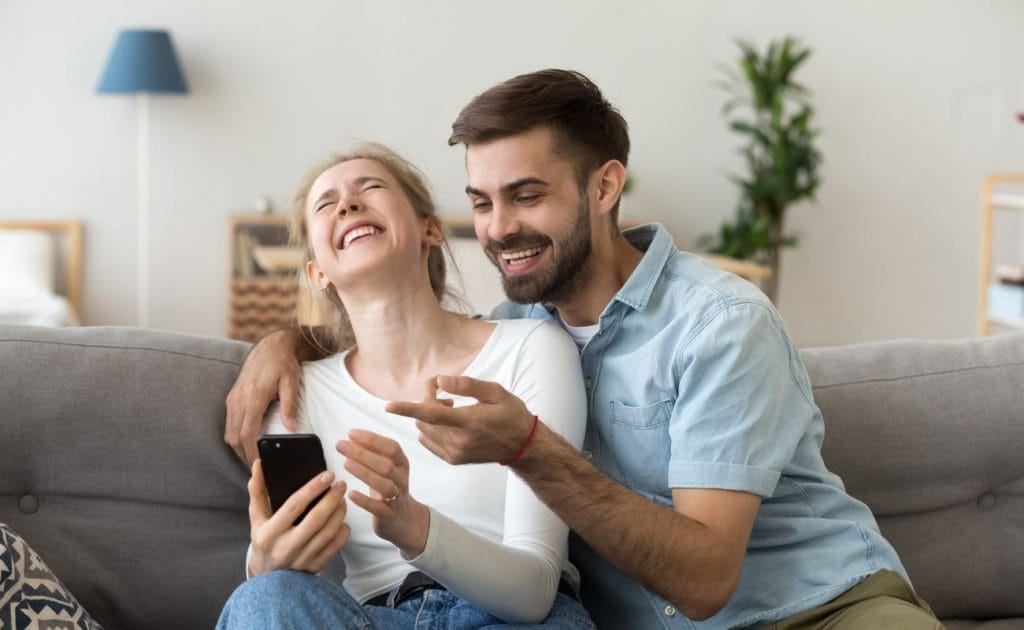 couple sitting together on the couch laughing about funny online joke holding a phone