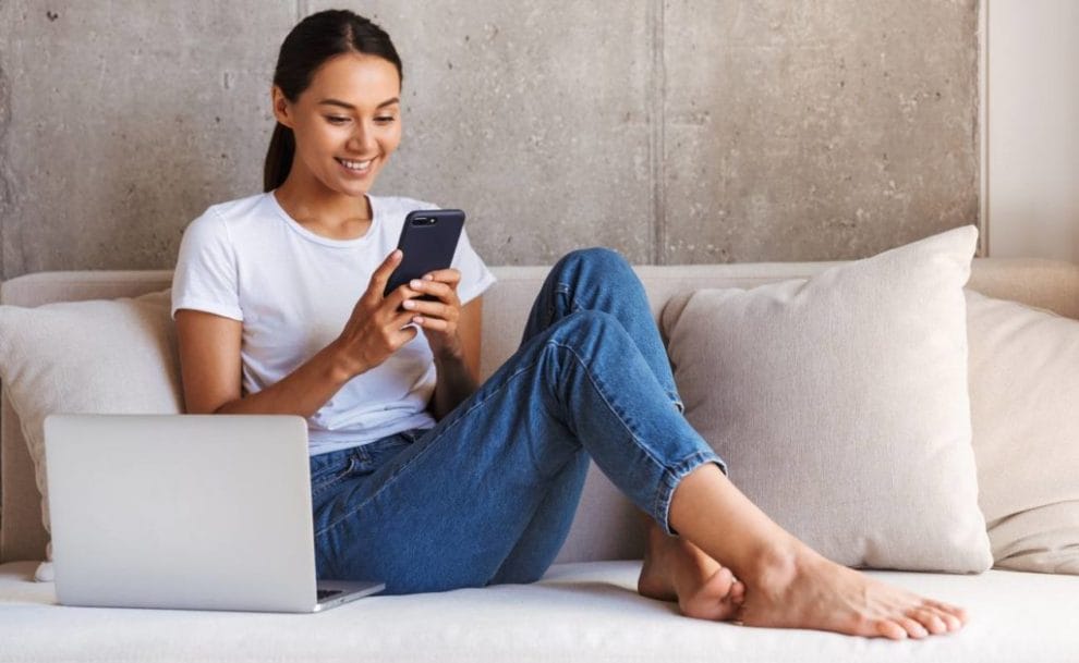 Smiling young woman using mobile phone while sitting on couch next to laptop