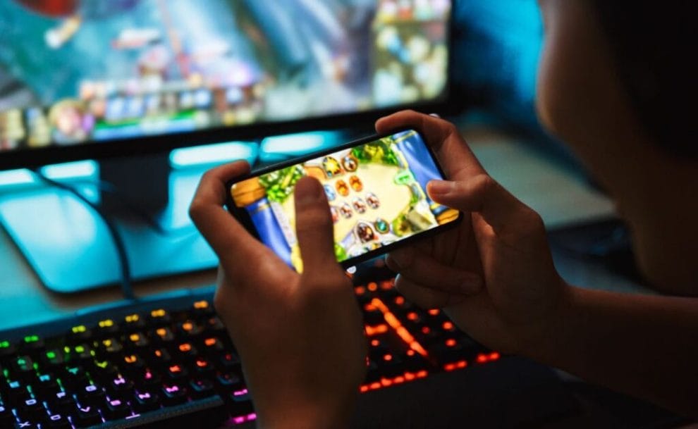 Gamer playing video games on smartphone and computer in dark room wearing headphones using backlit colorful keyboard