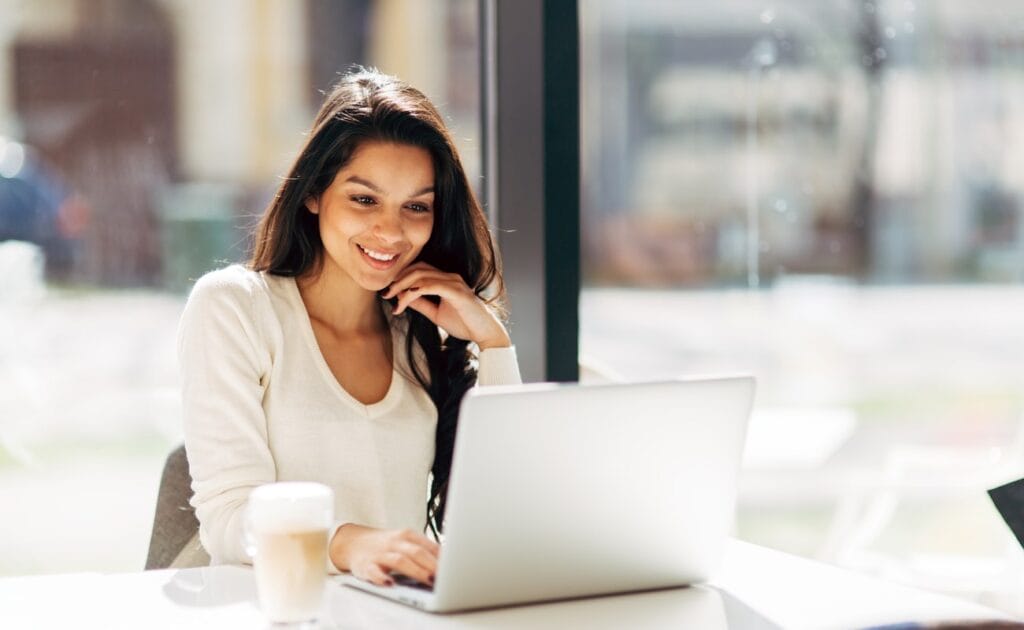 Young woman playing on laptop and drinking coffee in brightly lit cafe 