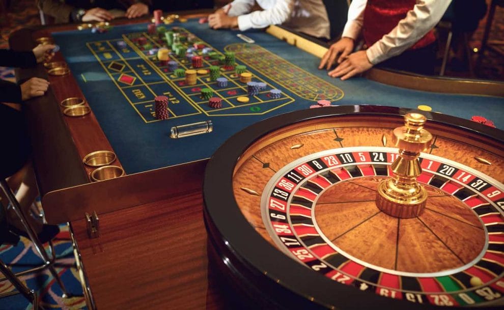 A roulette wheel and table in the background with croupier and players standing alongside