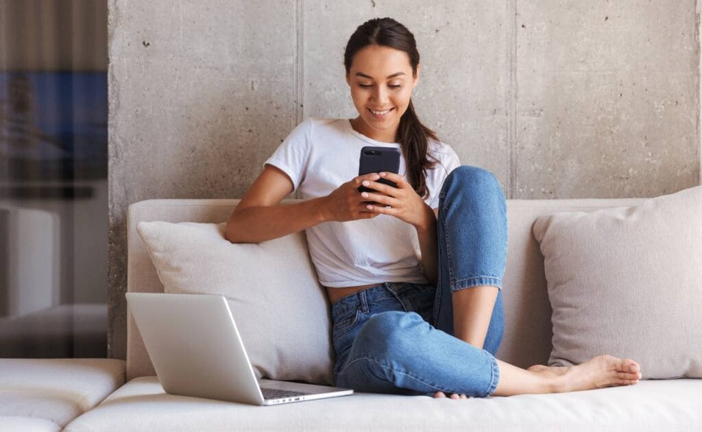 Woman using mobile phone while sitting a couch at home with laptop computer