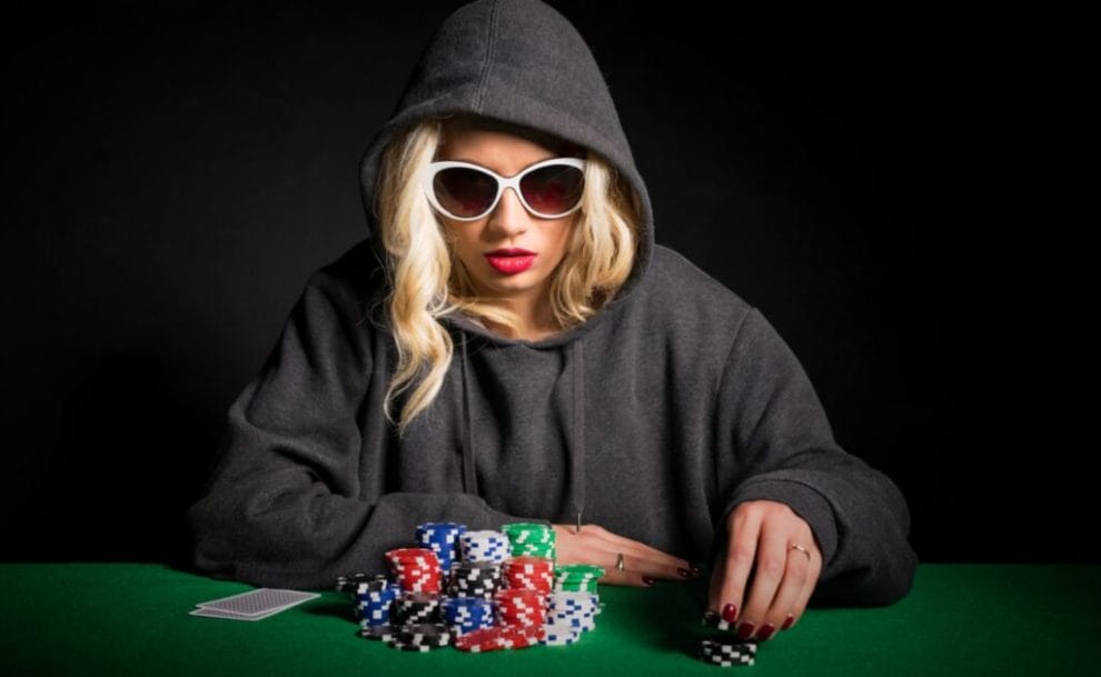 A woman wearing a dark hoodie and sunglasses sits at a poker table and reaches for her casino chips.