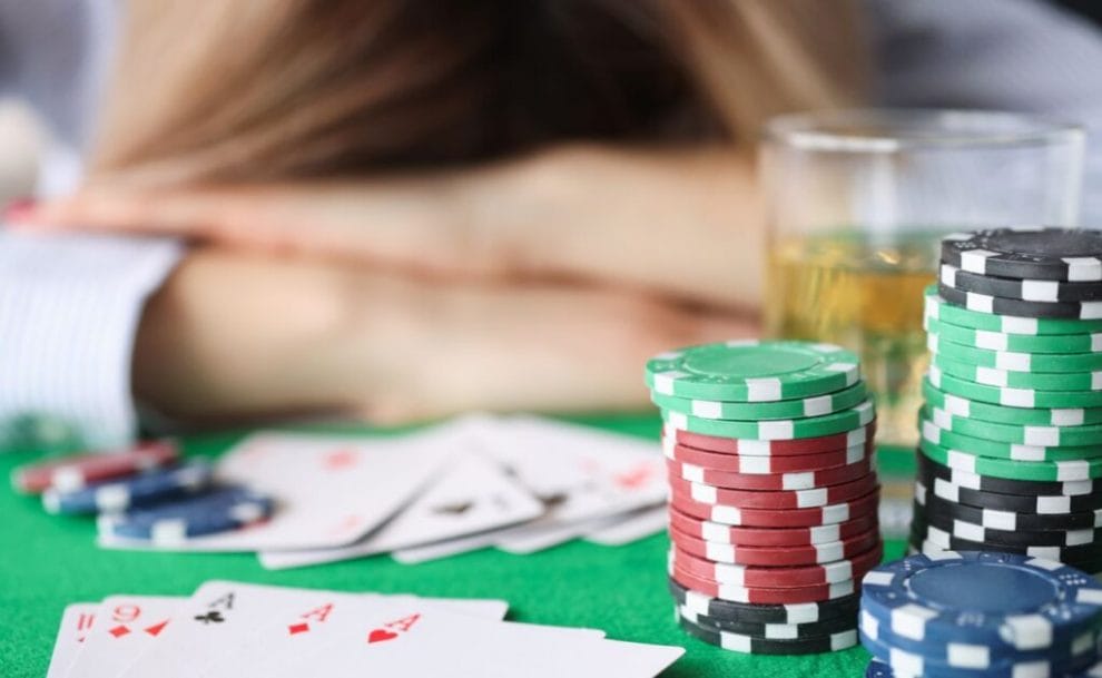 A woman lies on her arms on a casino table, an alcoholic drink and stacks of casino chips in front of her.