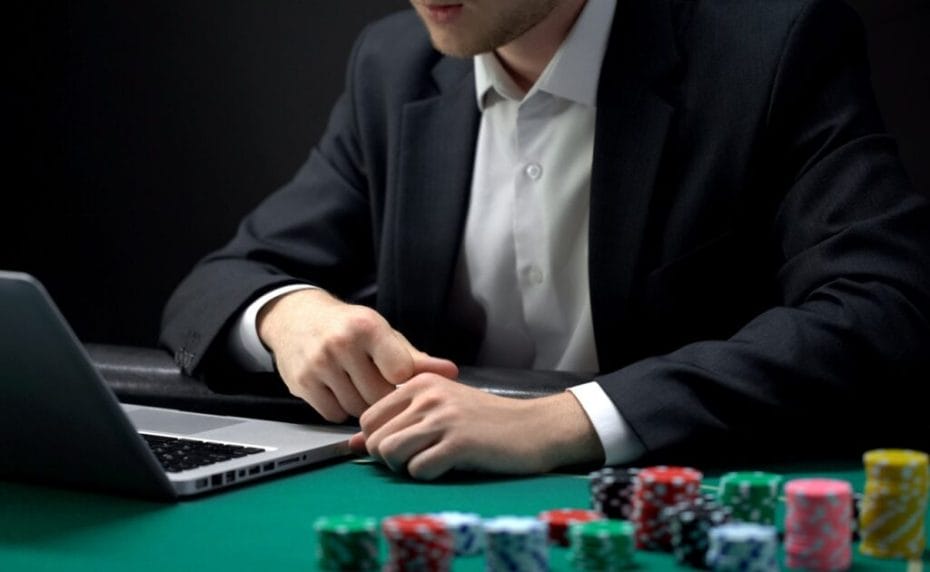 An online casino player wearing a suit is focused on his laptop. A stack of casino chips sits to his left. 