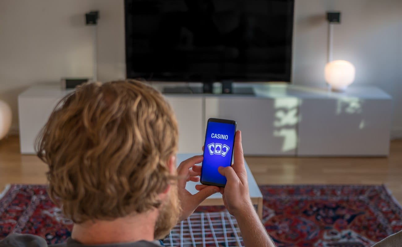 A man plays online casino games on his phone while sitting in front of a big TV.