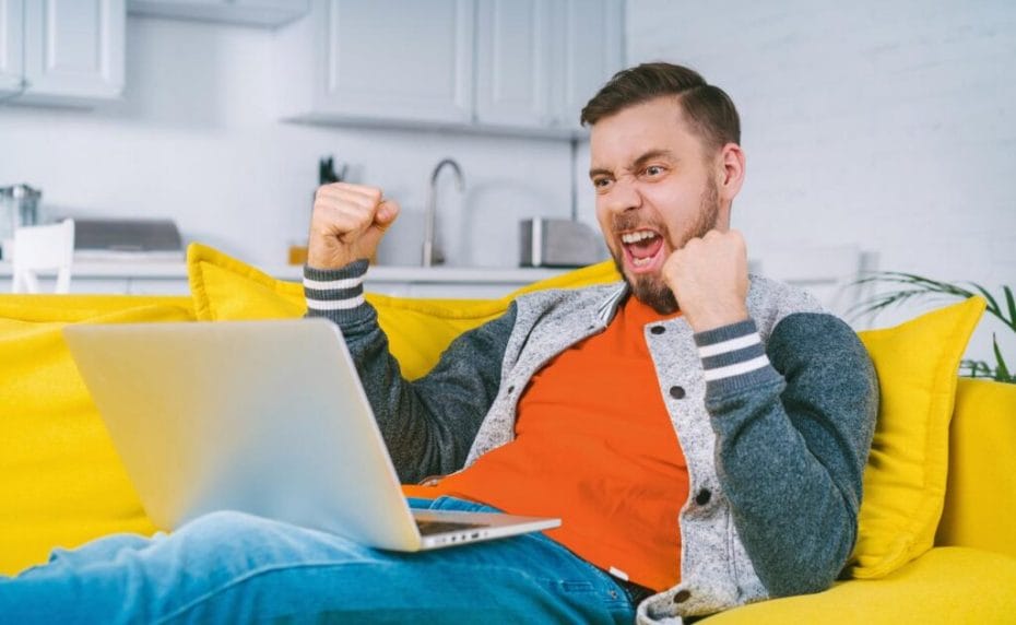 A young man celebrates a win at an online casino with a laptop on a yellow couch.
