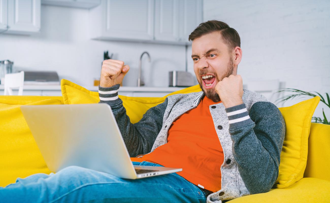A young man celebrates a win at an online casino with a laptop on a yellow couch.