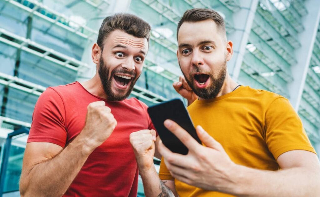 Two excited men celebrate an online gambling bet with a mobile device.