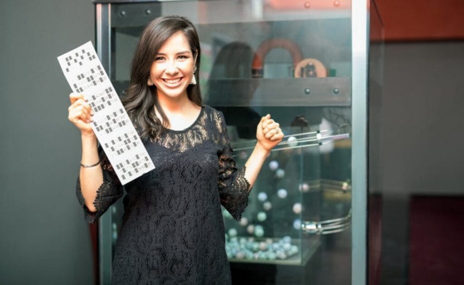 A young woman holds up her bingo card in front of a bingo ball machine.