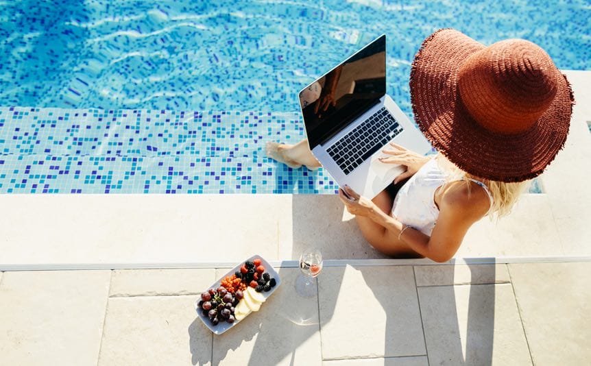 Woman using a laptop working remotely near a swimming pool.
