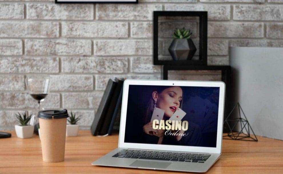 Laptop on a desk with an image of a casino online on the screen with a coffee cup and wine glass in the background.