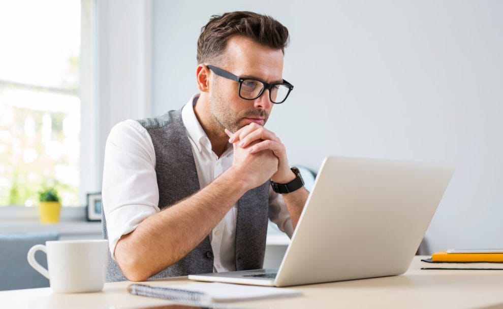 A man sits thinking in front of his laptop computer.