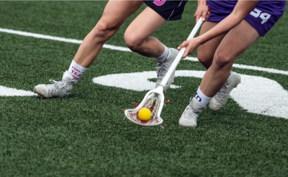 A lacrosse player on a field scoops the ball from an opponent and carries it forward.