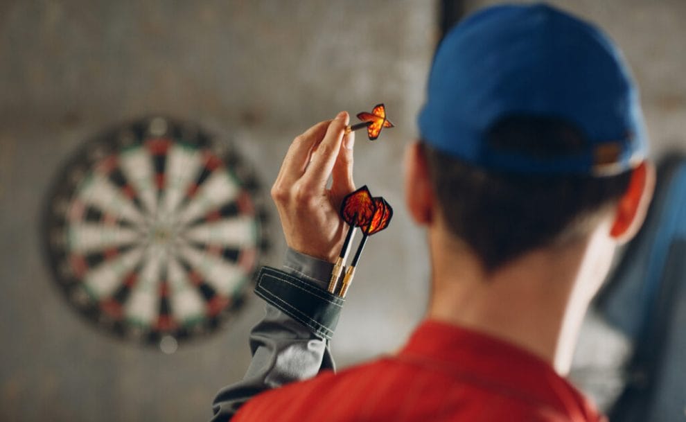 Darts player in a blue cap aims a dart at a dartboard.