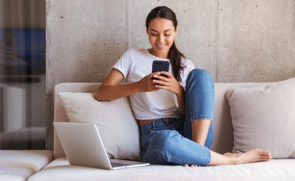 Happy woman using mobile phone while sitting on a couch at home with laptop computer.