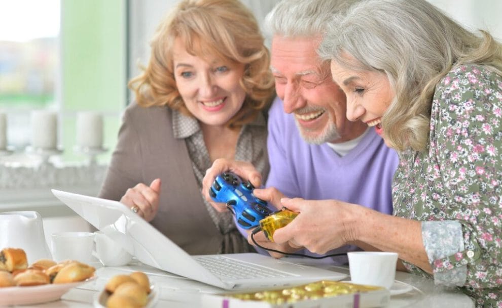 Older man and two women gaming on a laptop while drinking tea.