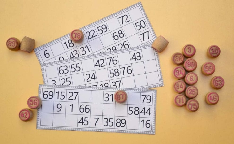 Three 90-ball bingo tickets on a beige surface surrounded by numbered wooden markers.