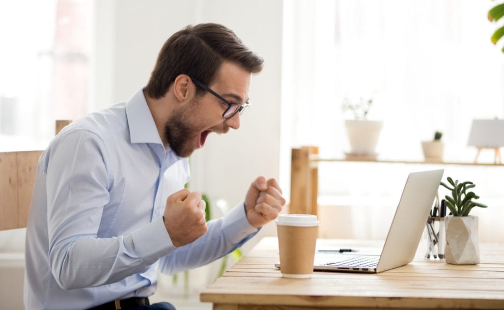 A euphoric man clenches his fists in front of his laptop.