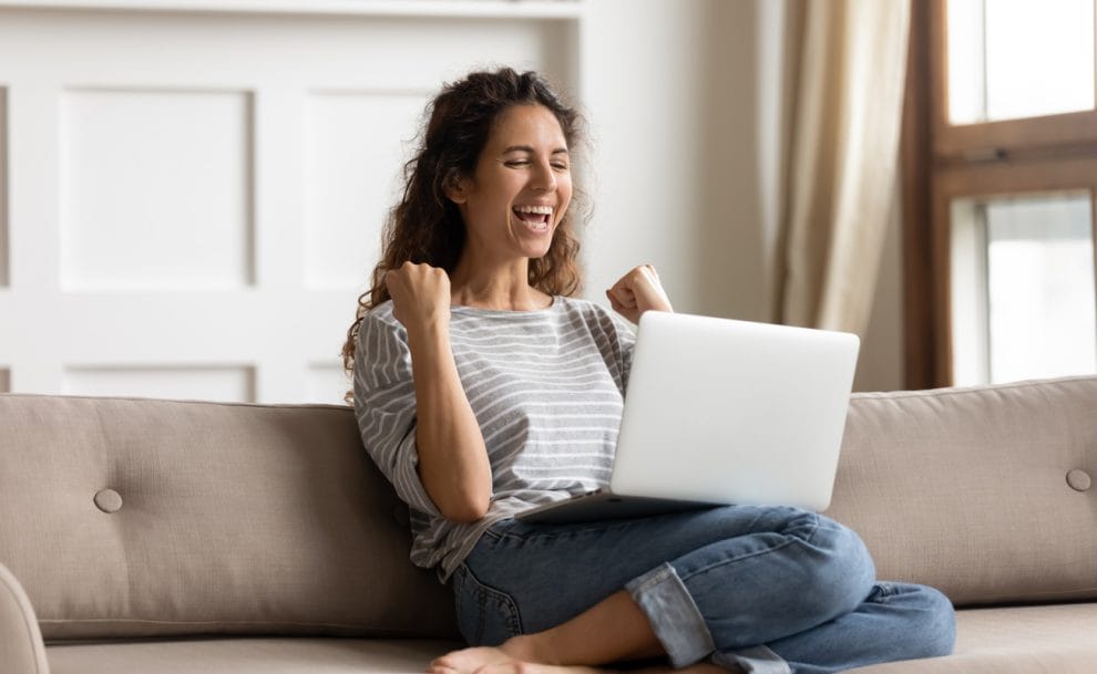 A woman on a couch pumps her fists in victory while playing casino games on a laptop.