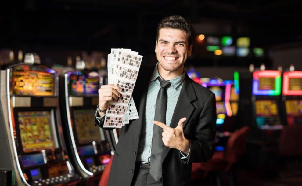 A smiling man in a suit holds up winning bingo cards with slot machines in the background.
