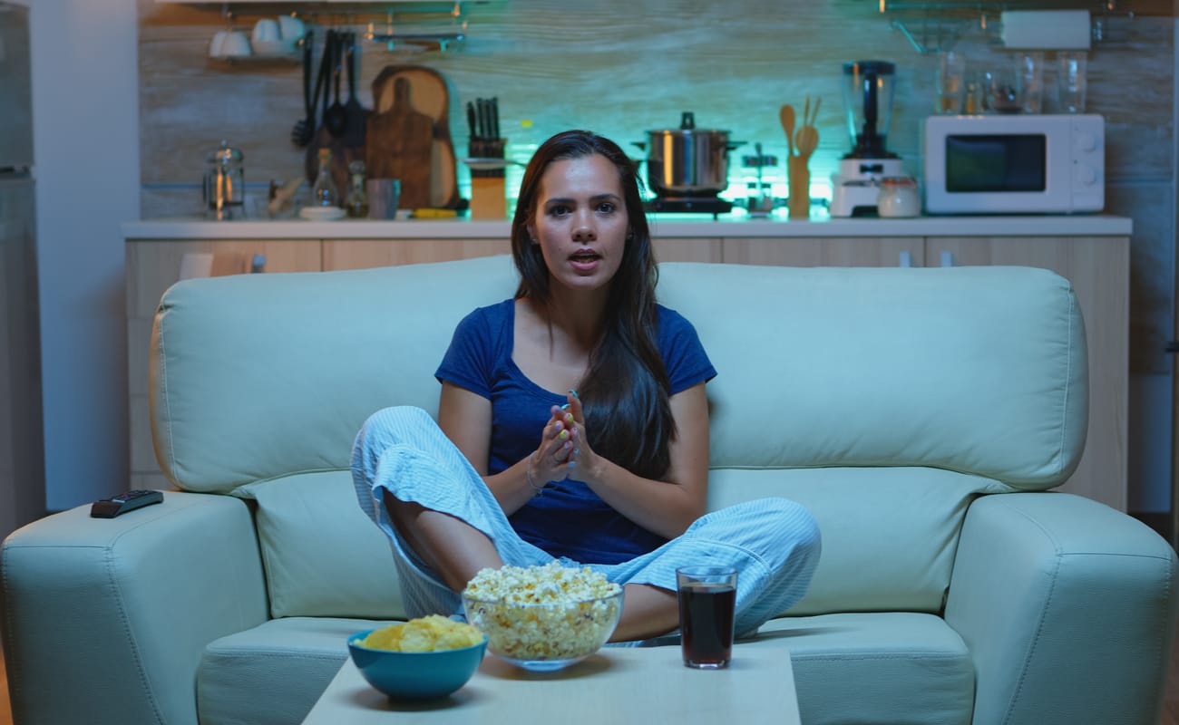 An eager looking woman sitting on the couch with snacks and a drink waiting to watch TV.