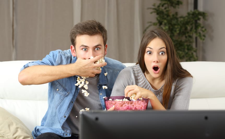 A couple looks amazed watching TV and eating popcorn.