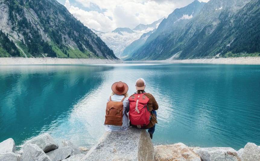 A couple with backpacks and hats sit in front of a lake.