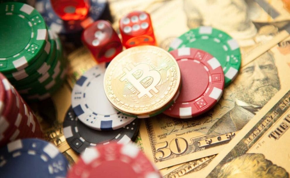A representation of a Bitcoin token sits on a pile of casino chips, surrounded by dice, money and chip stacks.