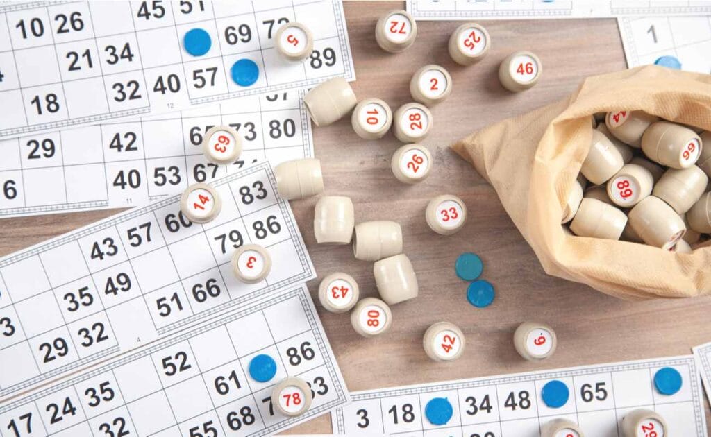 Table full of 90-ball bingo tickets, a bag full of wooden barrels with numbers on them and blue markers