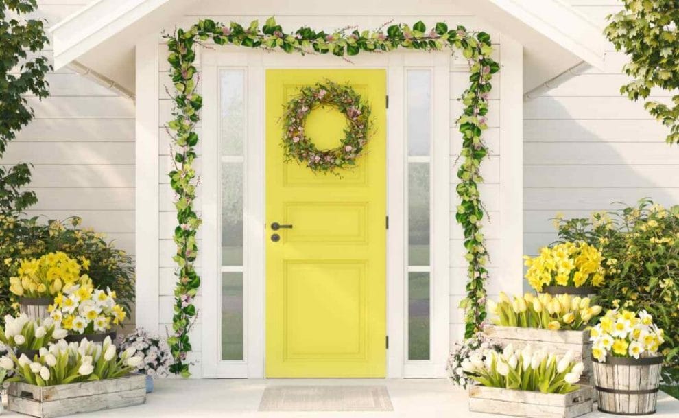 A porch with a yellow door and a wreath, surrounded by yellow flowers and green plants.