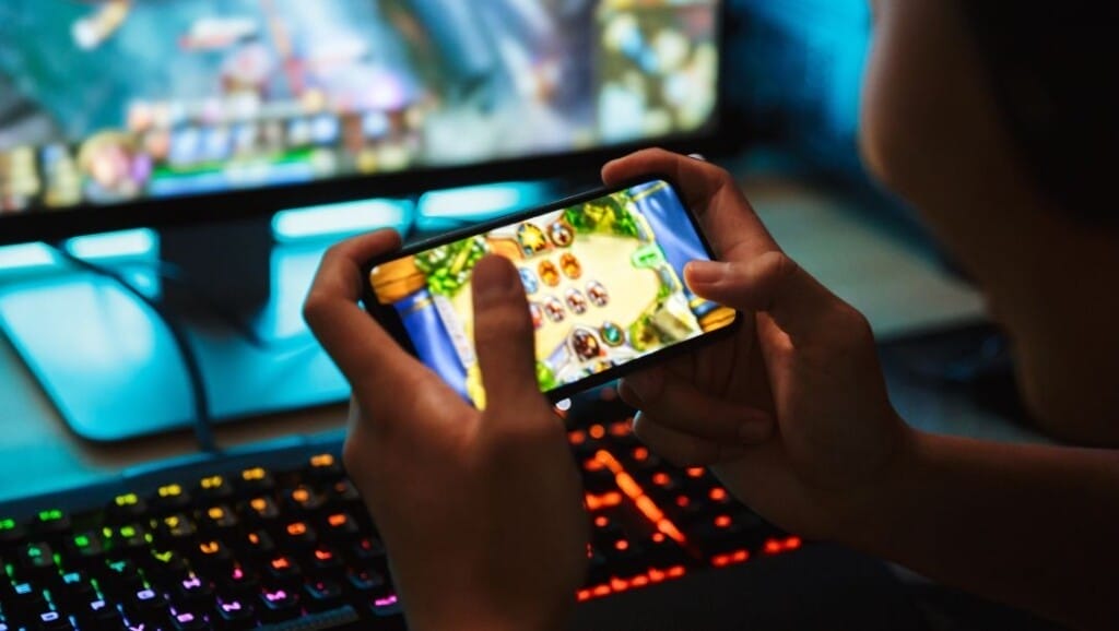 Person playing a colourful mobile game on a smartphone while sitting at a computer desk with a rainbow-lit mechanical keyboard and a gaming monitor in the background.