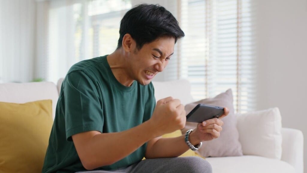 Man sitting on a sofa at home, clenching his fist in victory while playing an online casino game on his smartphone.