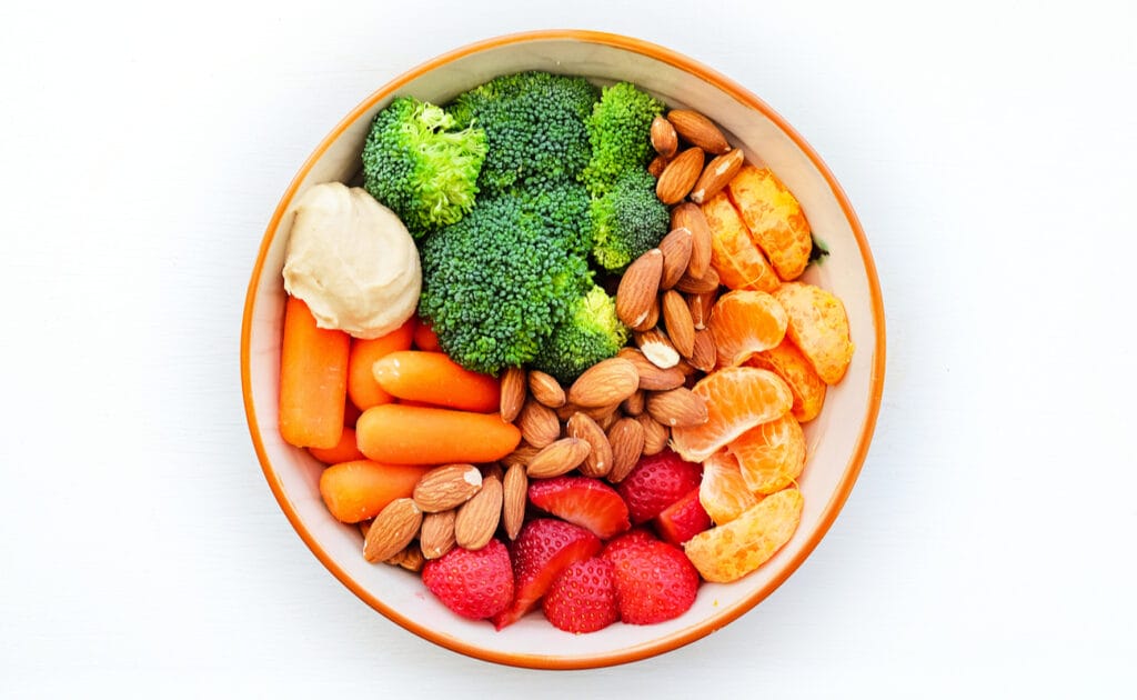 A bowl full of healthy snacks like nuts, strawberries, broccoli and carrots.