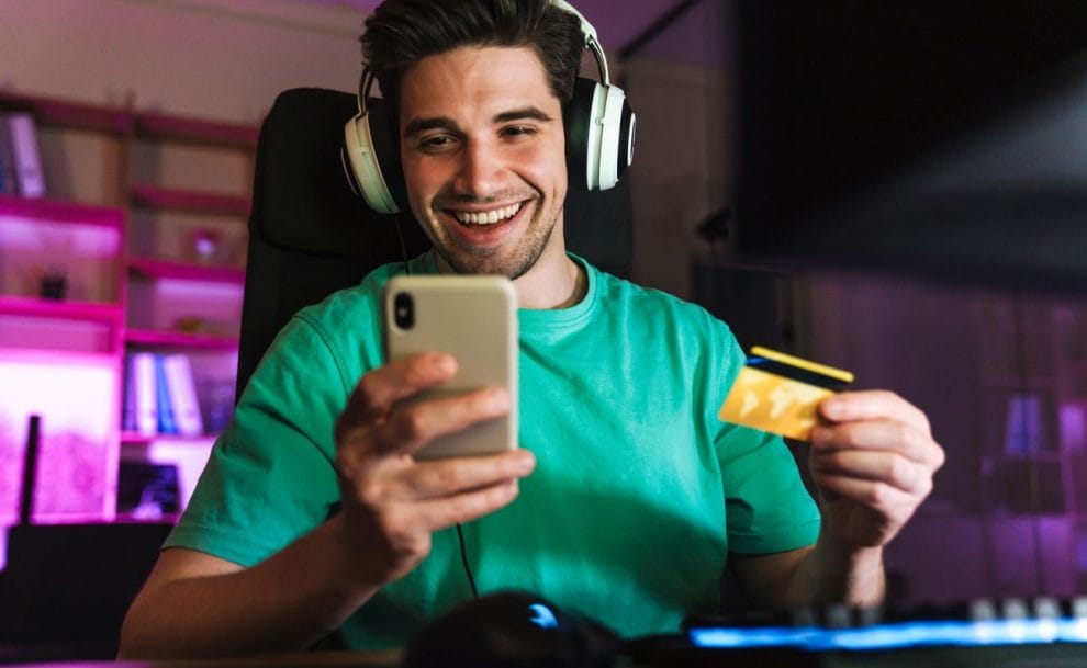 Man wearing a headset in front of a computer looking at his smartphone and holding a credit card.