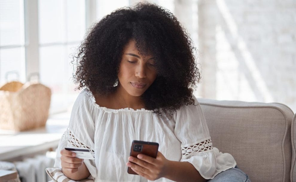 Woman sitting on a couch looking at a smartphone screen and holding a credit card.
