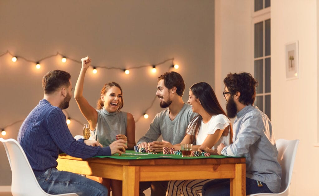 A group of friends sitting around a table with a board game and one woman with her hand in the air.