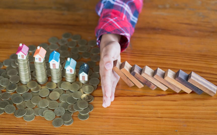 A hand stops a stack of dominoes from knocking over coins with houses on them.