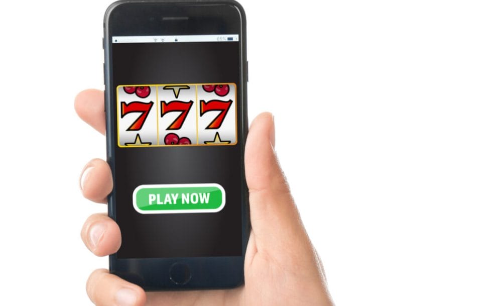A phone with a slot game and “Play Now” button, held in a person’s hand.