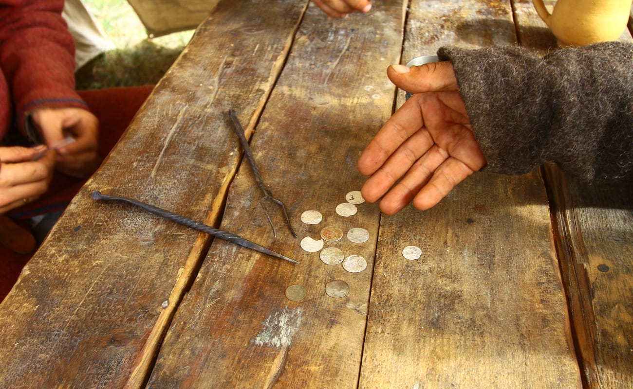 A man’s hand gestures to old-looking gold coins on a rustic wooden table.