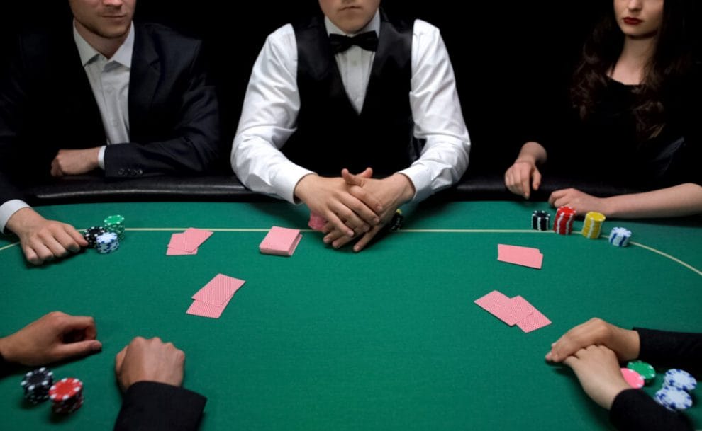 Four players and a dealer sitting around a poker table, each with their playing cards and poker chips in front of them.