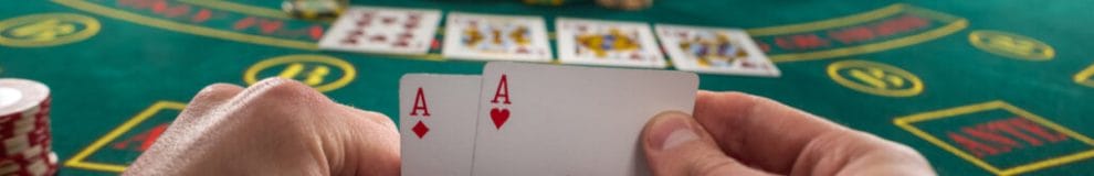 A poker player holds up two aces with the table and cards in the background.