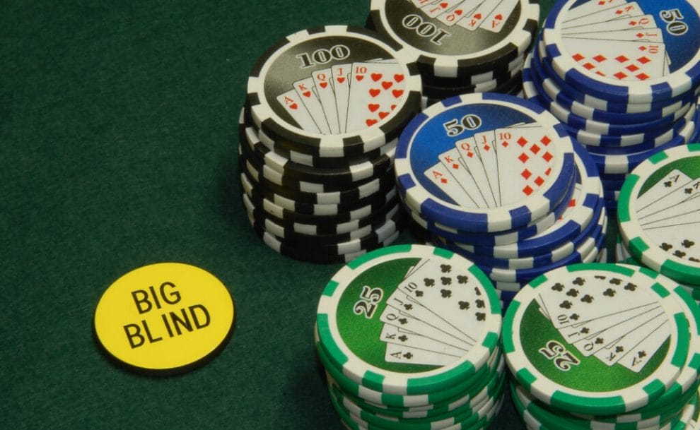 Stacks of green 25, blue 50, and black 100 poker chips and a single, yellow Big Blind chip lying on a green felt poker table.