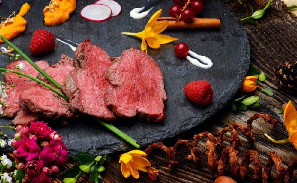 Slices of rare steak on a stone platter with berries, radishes, flowers, and other colorful foods.