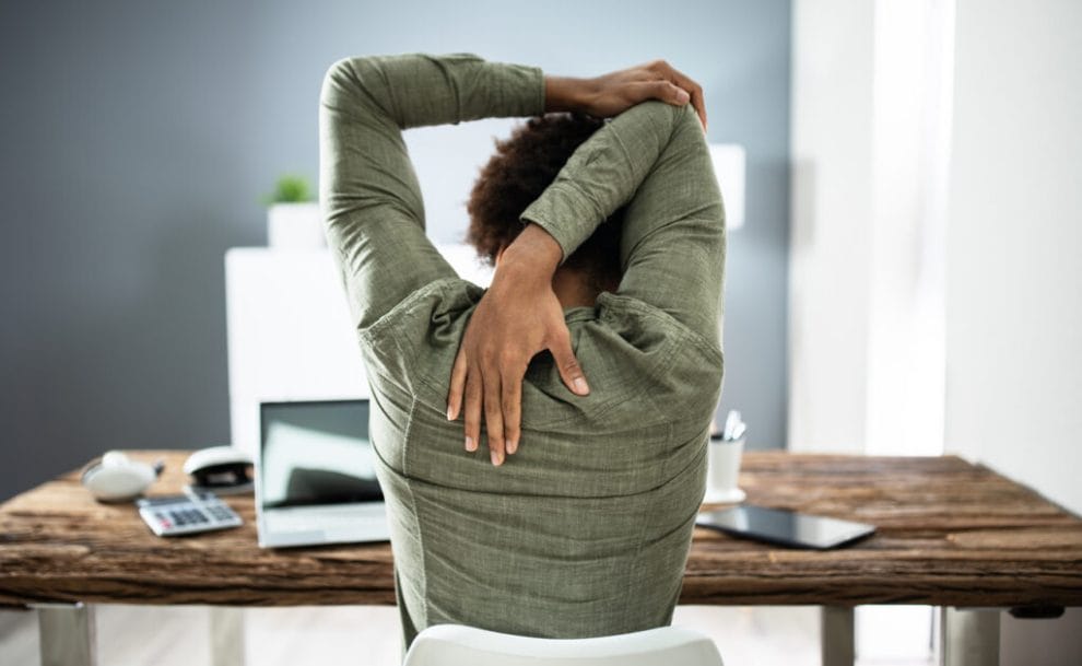 A view from the back of a woman doing a tricep stretch at her desk.