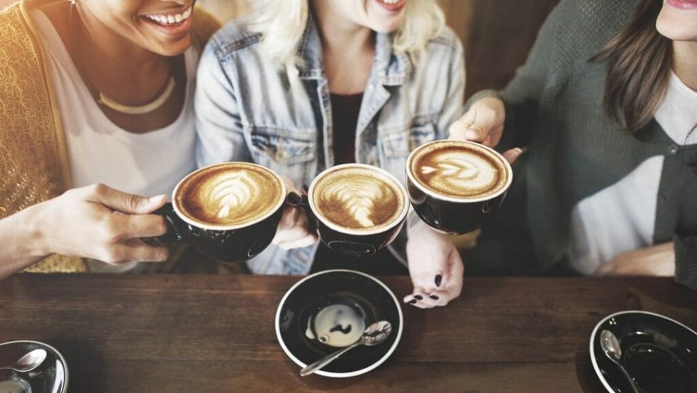 Women sitting at a table holding cups of coffee while smiling and laughing.