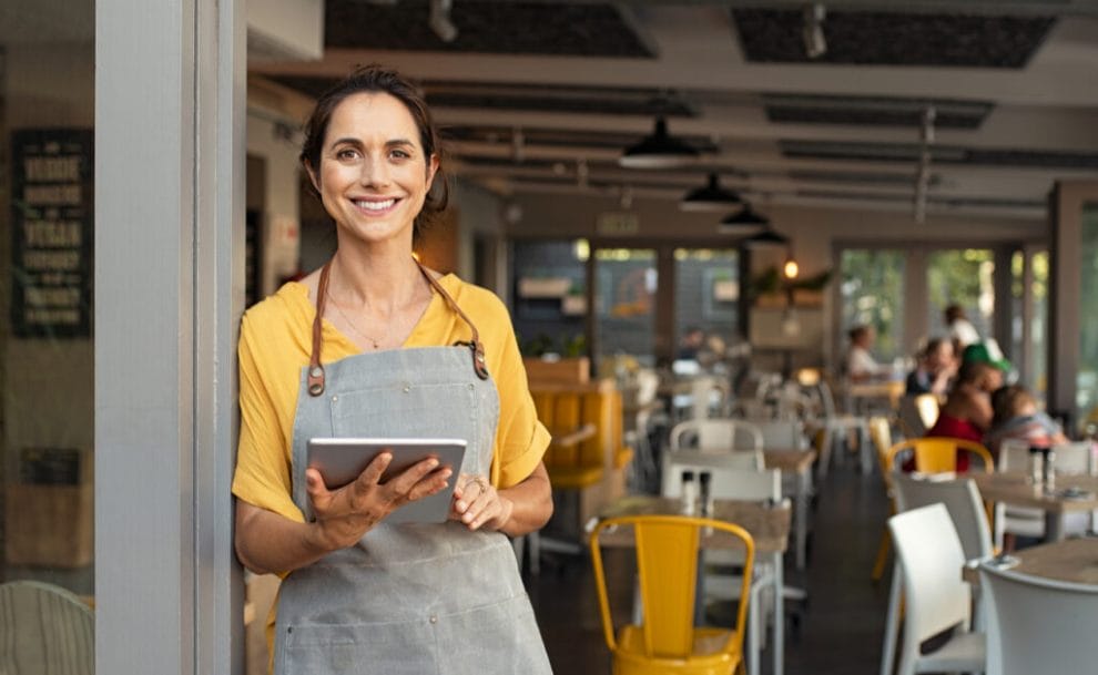 A smiling woman stands outside a restaurant with a tablet in her hand.